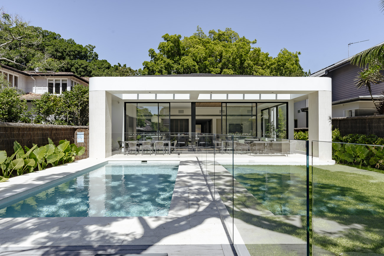 Frameless glass pool fence in front of a white pavilion and lawn at a Rose Bay residence