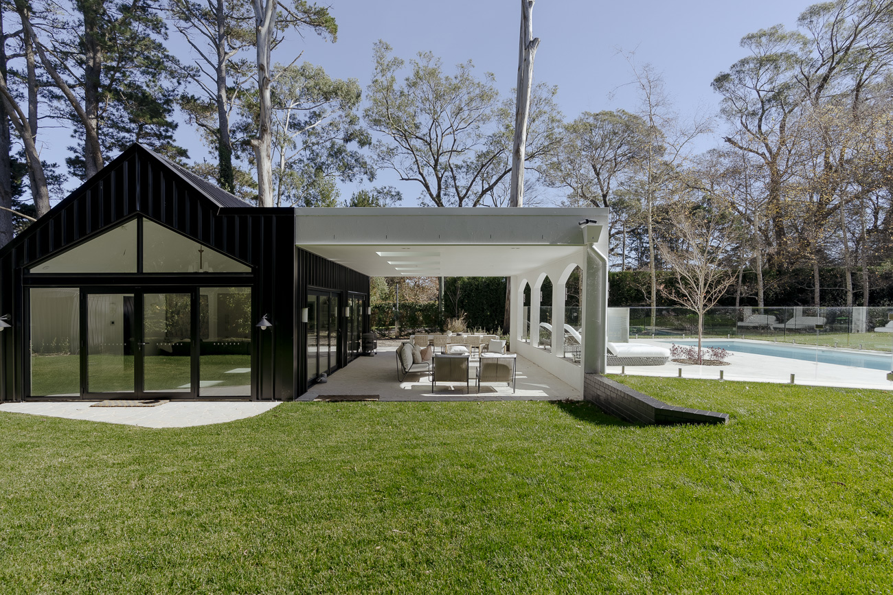 Frameless glass pool fence between a white arched pavilion and a black pool house on a lawn at a Burradoo residence