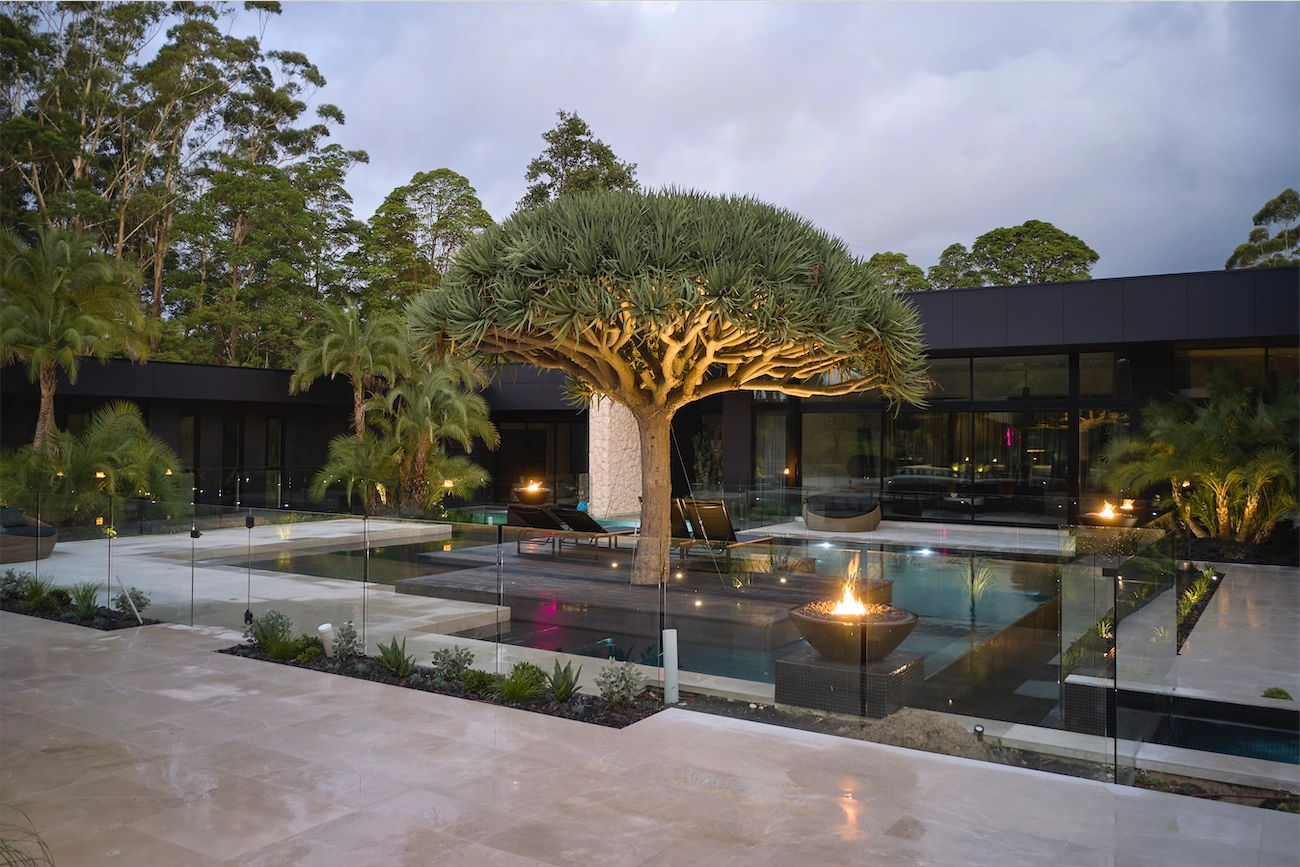 Frameless glass pool fence around a pool and lit fire pit at dusk at a Dural residence, beneath a dragon tree